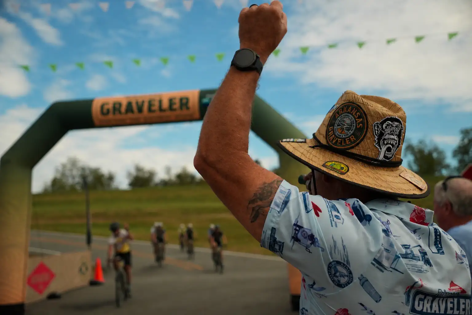 A Graveler in a patched straw hat raising a fist as riders reach the finish arch