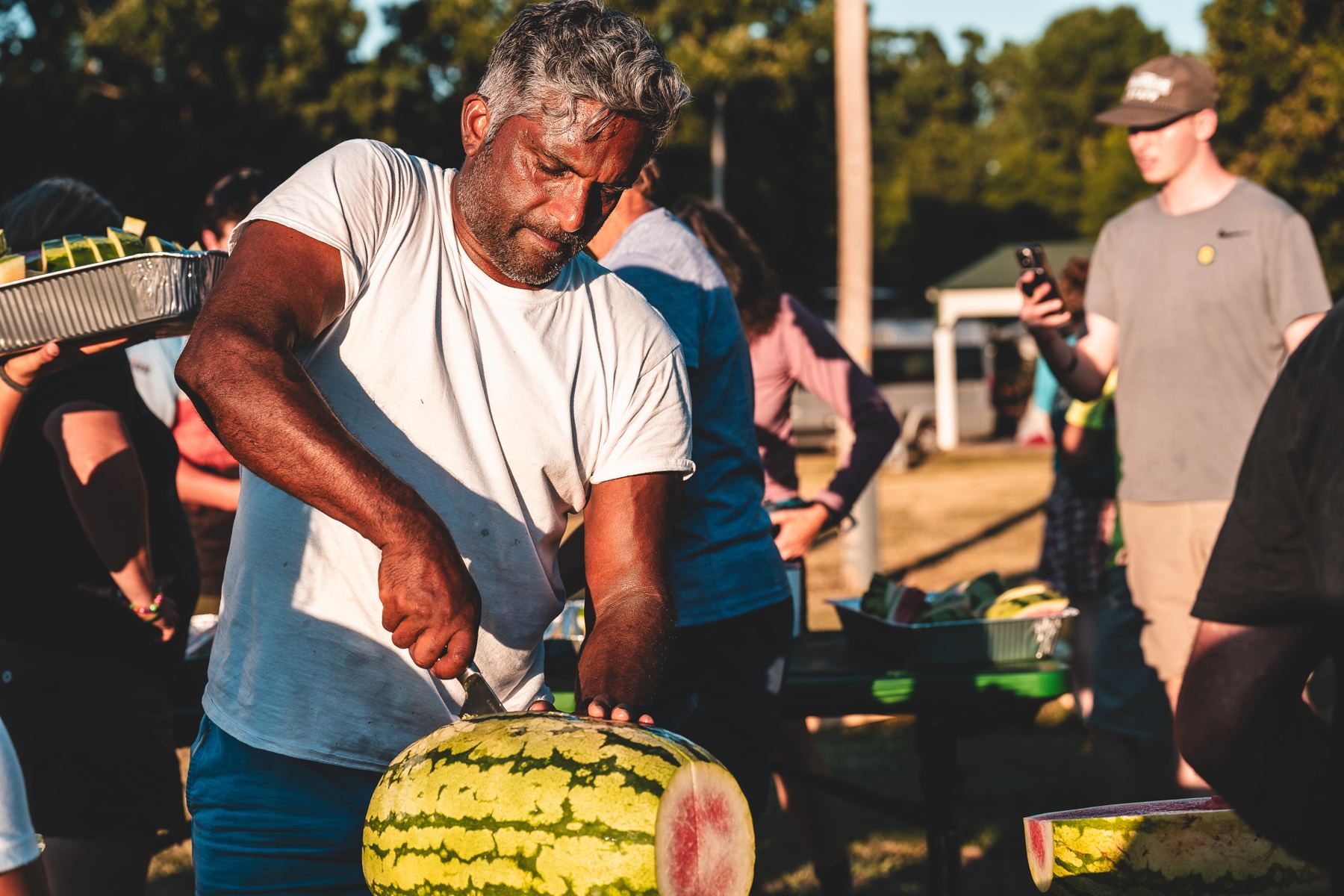 A man cutting watermelon for the camp in evening light while people gather around