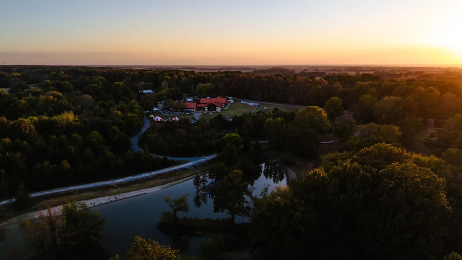 An aerial sunset view over the Day 2 finish: red-roofed lodges, striped event tents and a still pond ringed by Delta pines.
