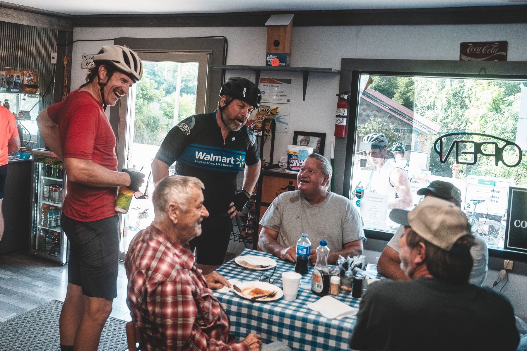 Riders and local men talking around a checked tablecloth inside a general store